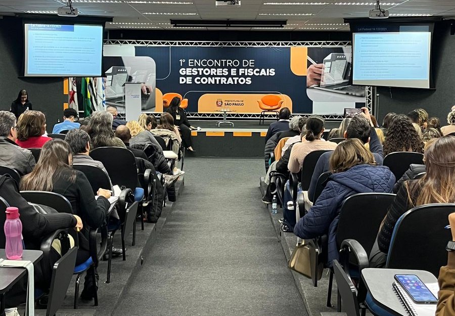 Imagem do auditório do evento, há um corredor ao centro e fileiras de cadeiras na esquerda e direita, onde há pessoas sentadas.