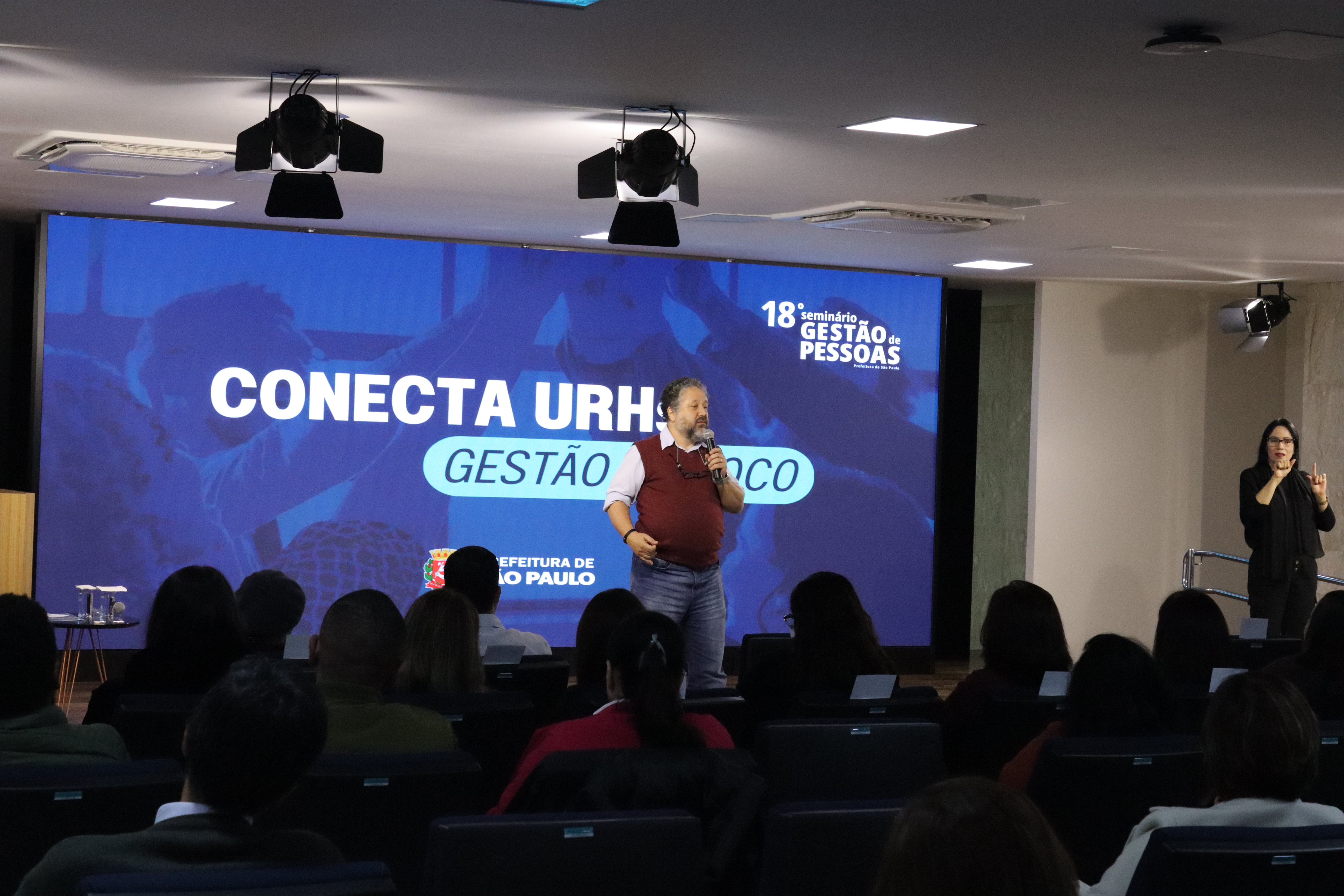 Imagem do auditório do décimo oitavo Seminário de Gestão de Pessoas. Ao fundo vemos um palestrante em frente a um telão com a logo do evento. A frente, vemos o perfil do público de costas.