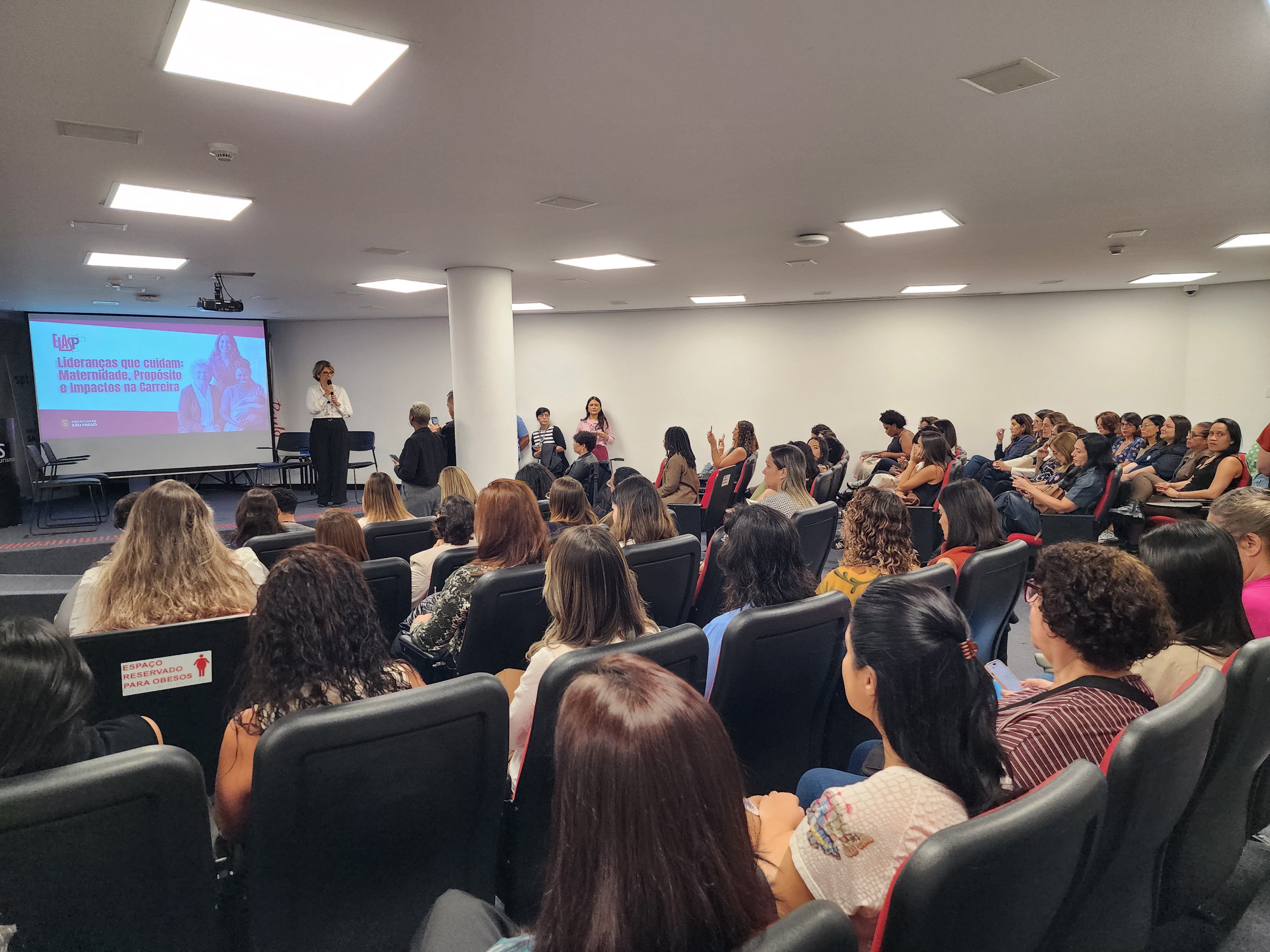 Imagem do auditório do evento, com a plateia de costas asssistindo a uma palestra.